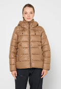 Frau trägt eine hellbraune, strukturierte Steppjacke mit Kapuze und schwarze Hosen, steht vor weißem Hintergrund.