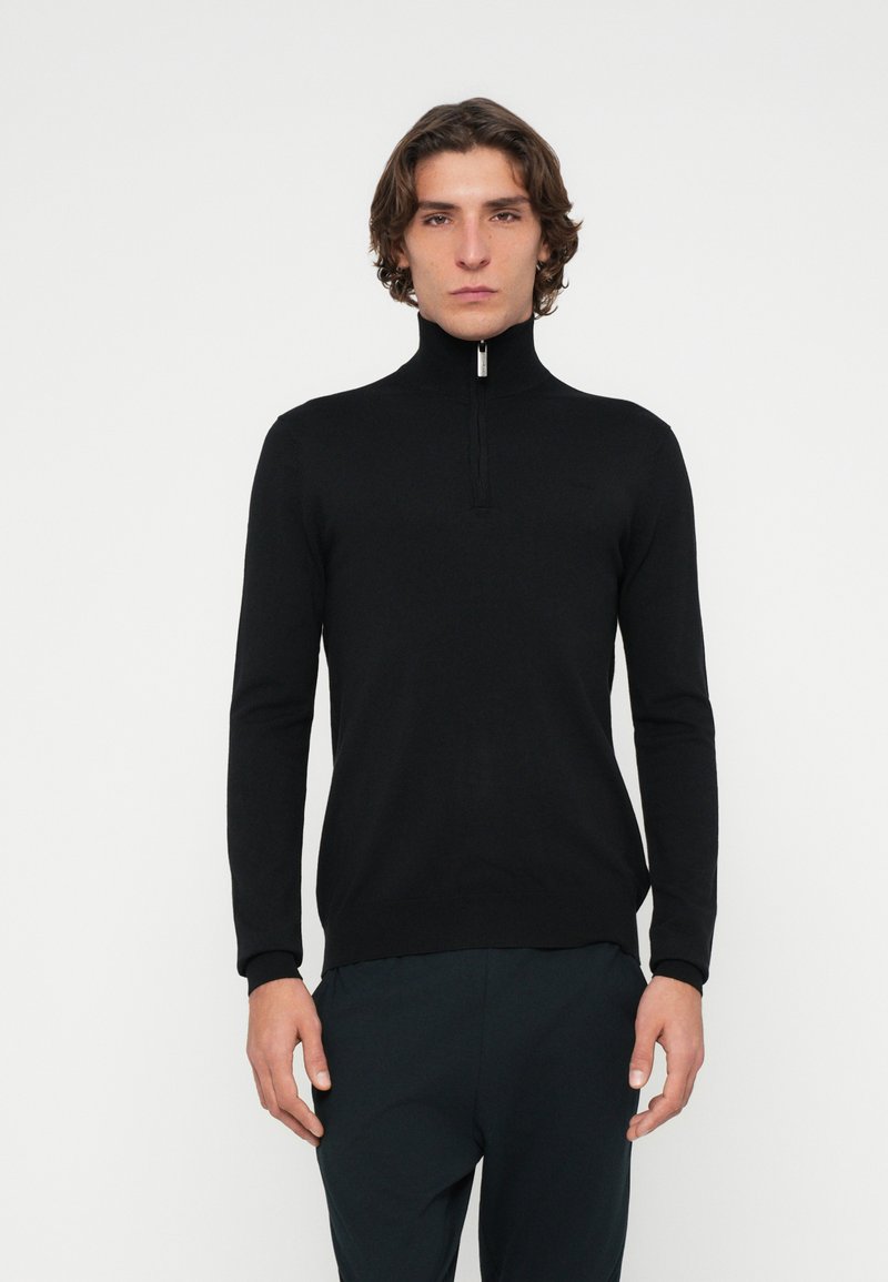 Schwarzer Pullover mit Reißverschluss am Kragen und langen Ärmeln, aus glattem Stoff gefertigt. Verfügt über einen hohen Kragen und minimale Details. Getragen mit dunkler Hose.