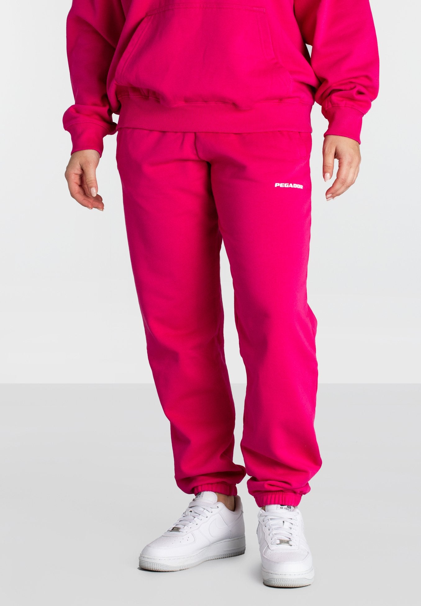 Person, die helle pinke Jogginghosen mit weißem "Pegador"-Logo und weißen Nike Air Force 1 Sneakern trägt, steht vor einem neutralen Hintergrund.
