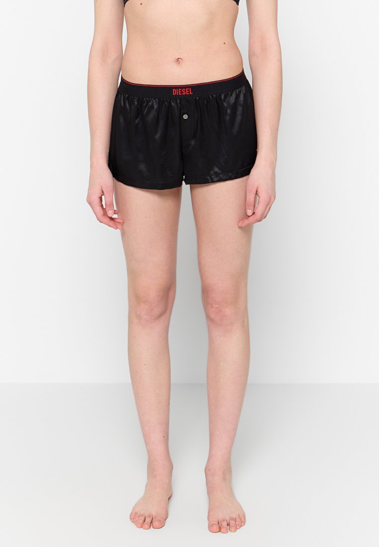 Frontalansicht einer Person, die schwarze Satin-Boxershorts von Diesel mit elastischem Bund und einer Einzelknopfdetails trägt und barfuß vor einer weißen Wand steht.