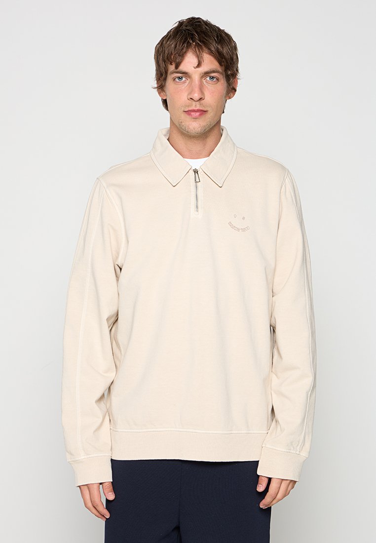 Mann trägt einen hellbeigen langärmeligen Pullover mit Reißverschlusskragen und dunkler Hose, steht vor einem schlichten hellen Hintergrund.