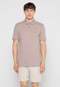 Mann trägt hell mauvefarbenes Poloshirt und beige Shorts und steht vor einem einfarbigen hellgrauen Hintergrund.