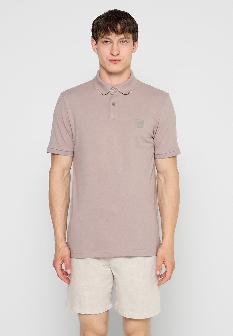 Mann trägt hell mauvefarbenes Poloshirt und beige Shorts und steht vor einem einfarbigen hellgrauen Hintergrund.