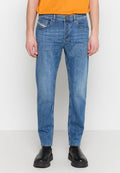 Mann in ausgewaschenen blauen Jeans, schwarzen klobigen Stiefeln und einem orangefarbenen T-Shirt, vor einem schlichten weißen Hintergrund stehend.