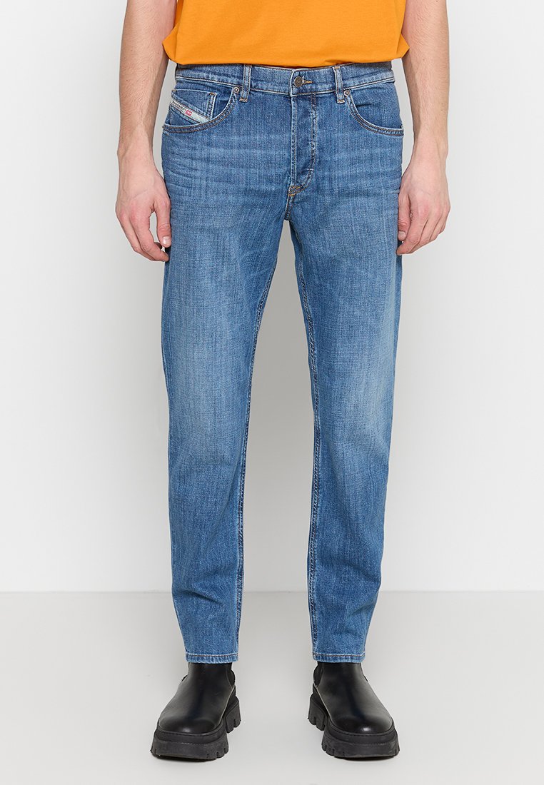 Mann in ausgewaschenen blauen Jeans, schwarzen klobigen Stiefeln und einem orangefarbenen T-Shirt, vor einem schlichten weißen Hintergrund stehend.