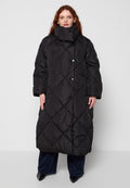 Frau mit langem roten Haar, die einen langen schwarzen Stepp-Puffer-Mantel mit hohem Kragen und schwarze Hose trägt und vor grauem Hintergrund steht.