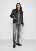 Schwarze Bomberjacke, graues Polohemd, hellgraue Jeans und schwarze Schuhe. Die Haare sind lang und wellig. Glatte Texturen und schlankes Design.