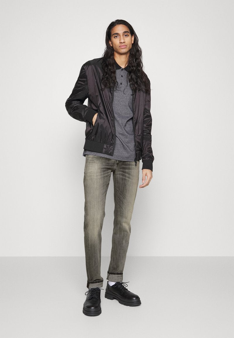 Schwarze Bomberjacke, graues Polohemd, hellgraue Jeans und schwarze Schuhe. Die Haare sind lang und wellig. Glatte Texturen und schlankes Design.