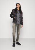 Schwarze Bomberjacke, graues Polohemd, hellgraue Jeans und schwarze Schuhe. Die Haare sind lang und wellig. Glatte Texturen und schlankes Design.