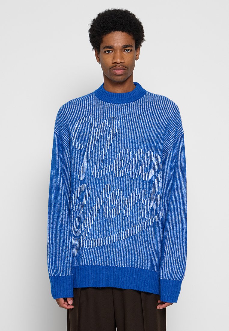 Junger Mann mit kurzen lockigen Haaren, der einen blau-weißen gestreiften Pullover mit dem Text "View of Beast" trägt, steht vor einem schlichten hellen Hintergrund.