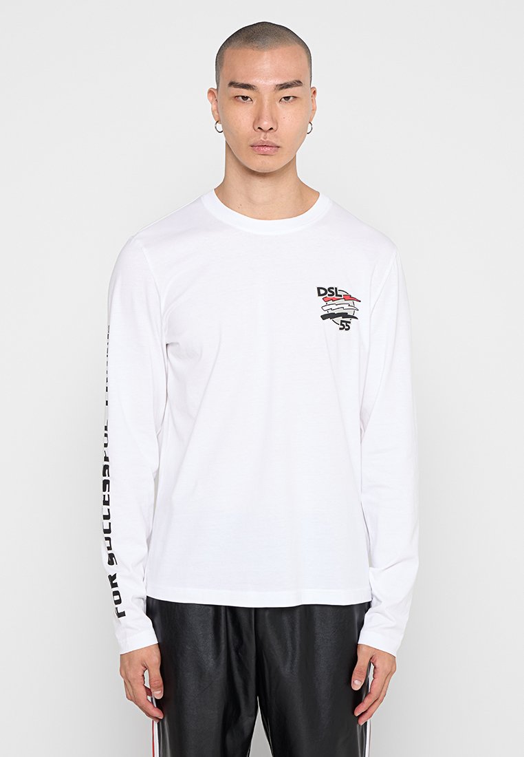 Junger Mann mit kurzen Haaren, der ein weißes Langarmshirt mit schwarzem und rotem "DSL 55"-Logo auf der Brust und Text am rechten Ärmel trägt.