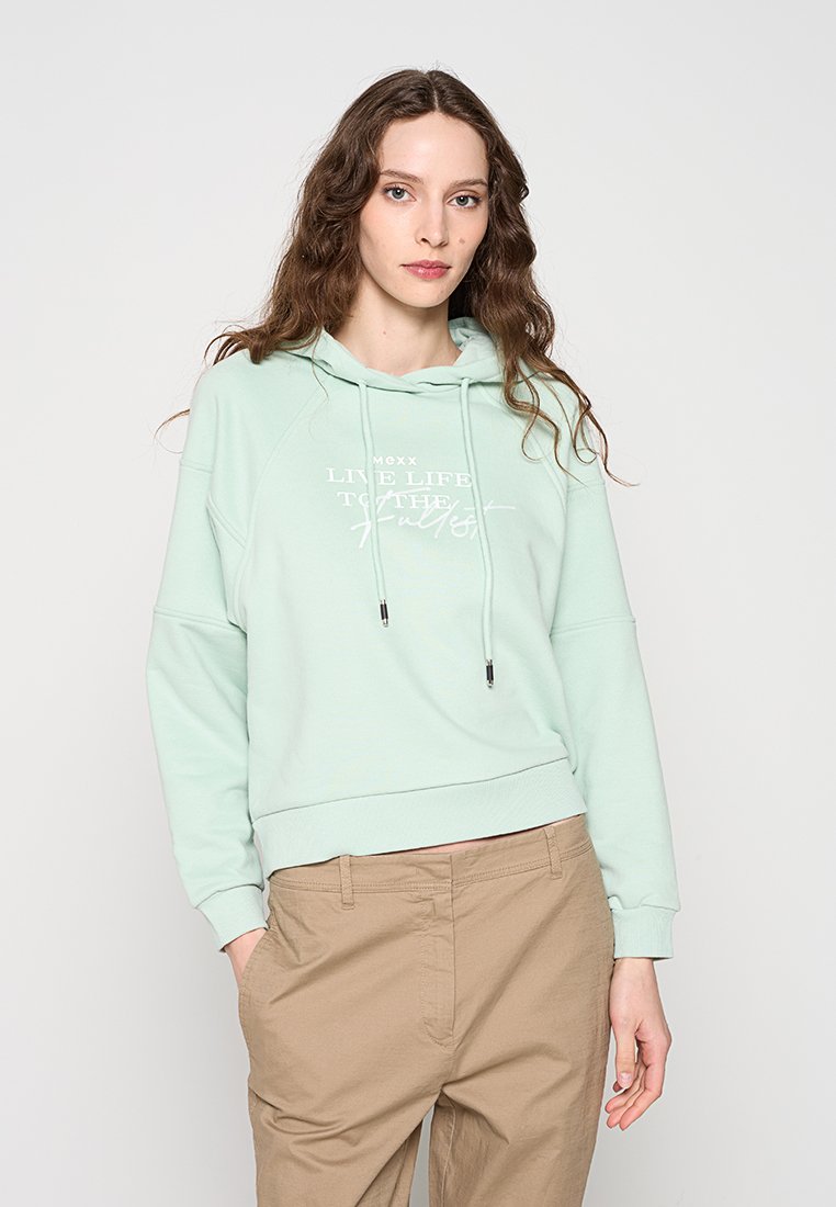 Junge Frau mit langen braunen Haaren trägt einen hellgrünen Hoodie mit weißem Schriftzug und beige Hose, steht mit einer Hand in der Tasche vor einfarbigem Hintergrund.