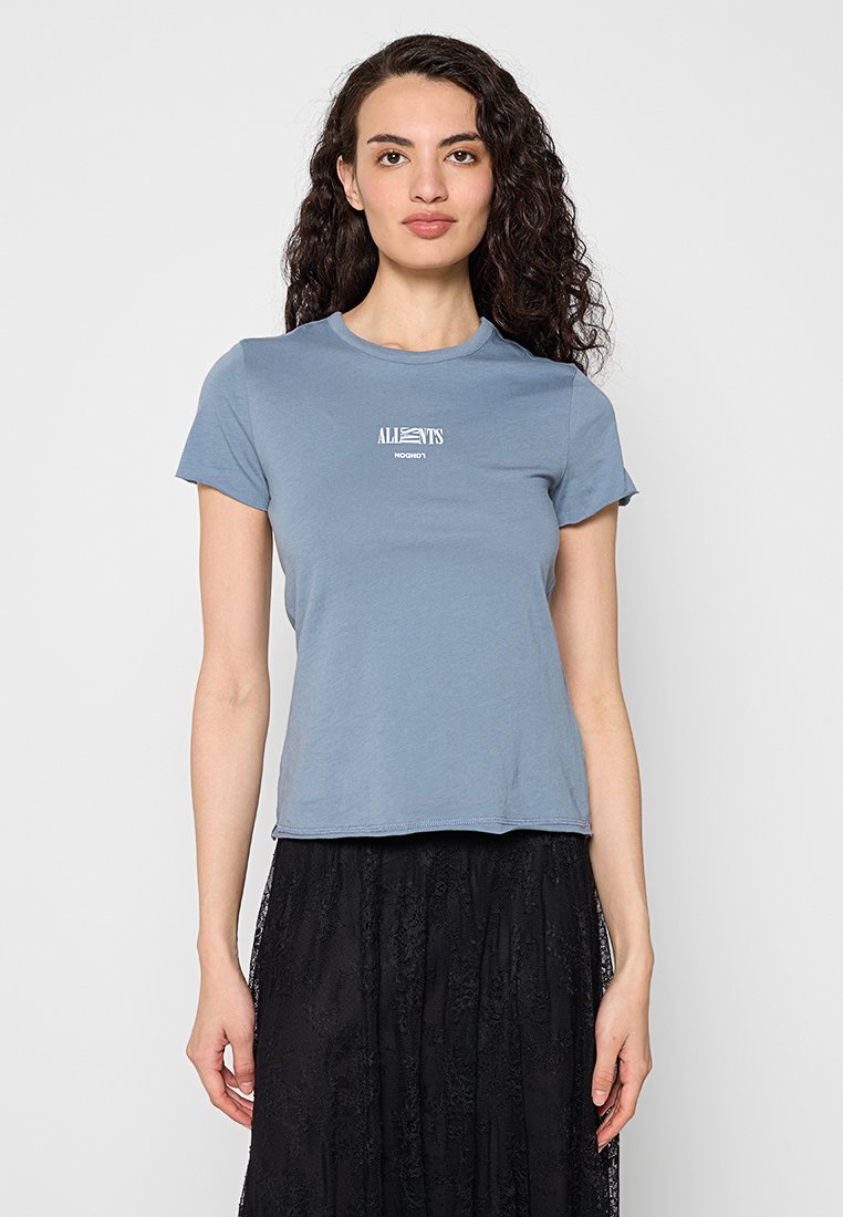 Frau mit lockigem Haar, die ein eng anliegendes blaues T-Shirt mit weißem Text und einen schwarzen Spitzenrock trägt, stehend vor einem schlichten hellen Hintergrund.