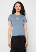 Frau mit lockigem Haar, die ein eng anliegendes blaues T-Shirt mit weißem Text und einen schwarzen Spitzenrock trägt, stehend vor einem schlichten hellen Hintergrund.