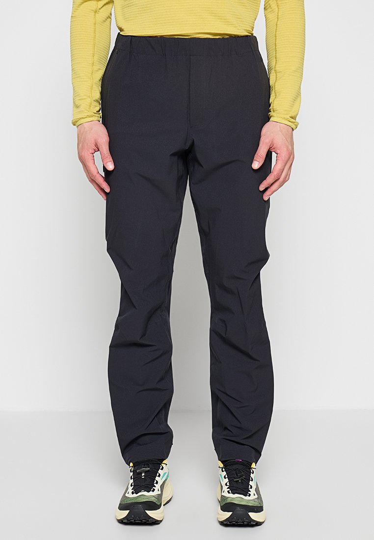 Person trägt schwarze elastische Bundhose, gelbes langärmeliges Shirt und mehrfarbige Turnschuhe, steht vor einfarbigem Hintergrund.