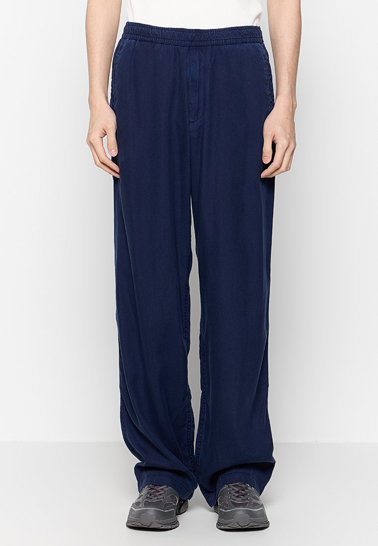 Person, der eine lockere dunkelblaue Hose mit elastischem Bund und grauen Turnschuhen trägt, vor einem schlichten weißen Hintergrund steht.