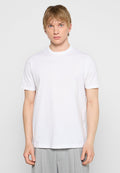 Junger Mann mit kurzen blonden Haaren, der ein einfaches weißes Diesel-T-Shirt und helle graue Hosen trägt, steht vor einem neutralen Hintergrund.