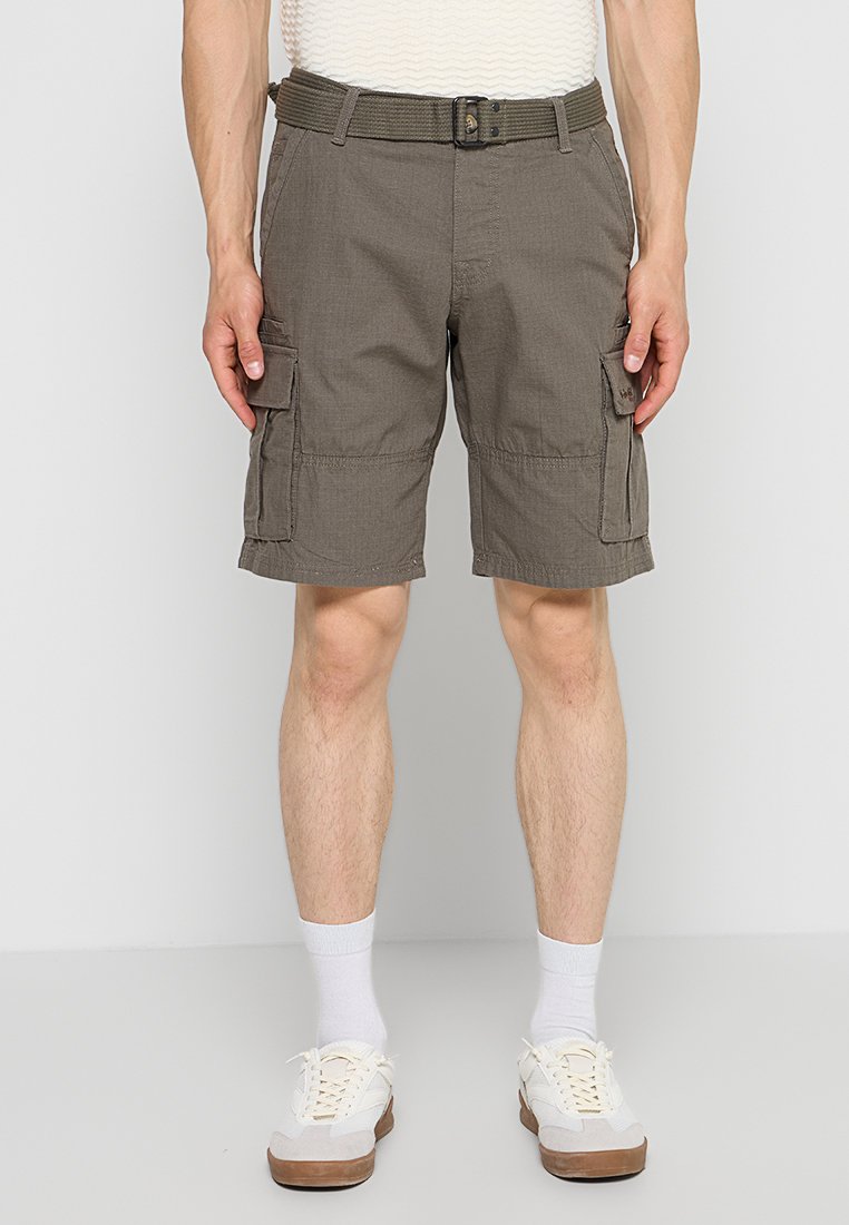 Mann trägt braune Cargoshorts mit Seitentaschen, weiße Socken und weiße Sneakers, steht vor einem schlichten hellgrauen Hintergrund.