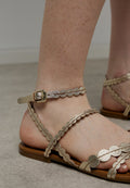 Goldfarbene flache Sandalen mit gewellten Riemen, verstellbarem Knöchelriemen und einer glatten braunen Sohle. Geeignet für warmes Wetter.