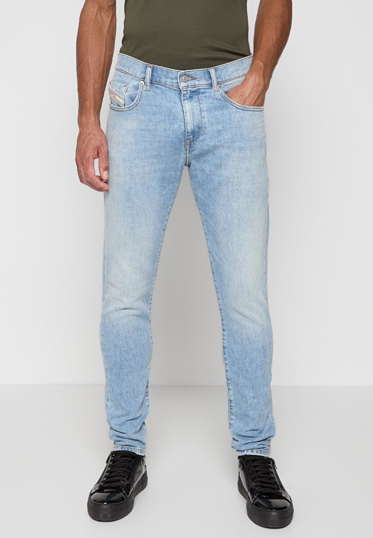 Mann in hellblauen Slim-Fit-Jeans, schwarzen glänzenden Sneakers und einem dunkelolivgrünen eng anliegenden Shirt, stehend vor einem einfarbigen grauen Hintergrund.