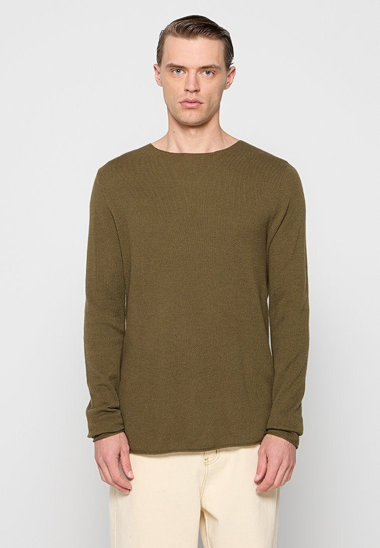 Mann in einem olivgrünen, strukturierten Langarmpullover und hellbeigen Hosen, der vor einem schlichten hellgrauen Hintergrund steht.