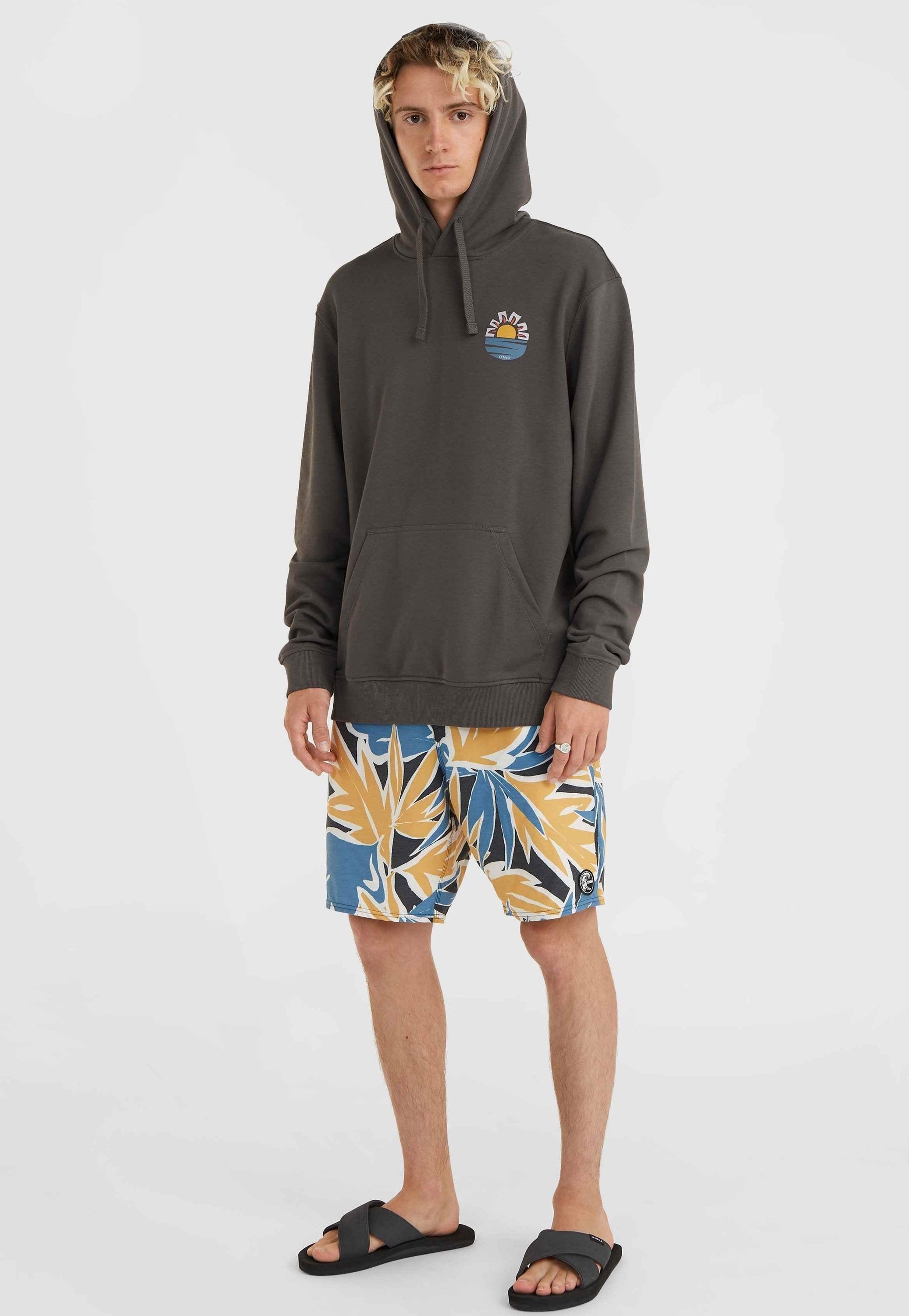 Dunkelgrauer Hoodie mit einer Fronttasche und einem bunten Sonnen- und Wellenmotiv. Kombiniert mit mehrfarbigen, floral gemusterten Badeshorts und schwarzen Sandalen.