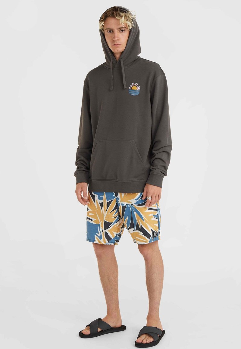 Dunkelgrauer Hoodie mit einer Fronttasche und einem bunten Sonnen- und Wellenmotiv. Kombiniert mit mehrfarbigen, floral gemusterten Badeshorts und schwarzen Sandalen.