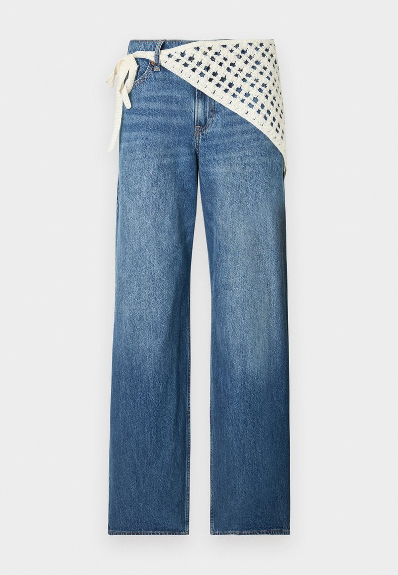Blaue Jeans mit weitem Bein und einem weißen Häkel-Wickeldetail, das auf Taillenhöhe auf einem einfarbigen Hintergrund gebunden ist.