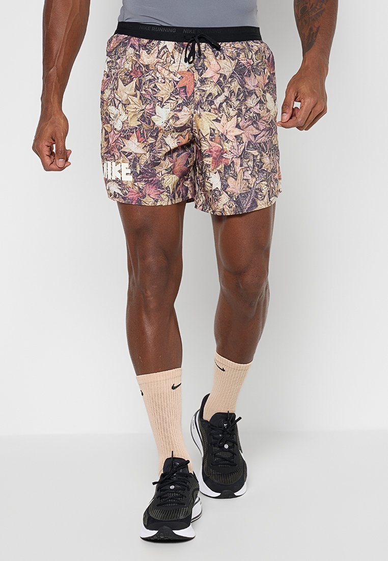 Mann trägt herbstlich gemusterte Nike-Shorts mit Blättern, beige Nike-Socken und schwarze Sneakers, steht vor einem schlichten grauen Hintergrund.