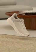 Hellbeiger Skechers-Sneaker mit dicker segmentierter Sohle und Mesh-Einsätzen, positioniert in der Nähe eines hölzernen Couchtischs mit gestapelten Büchern.