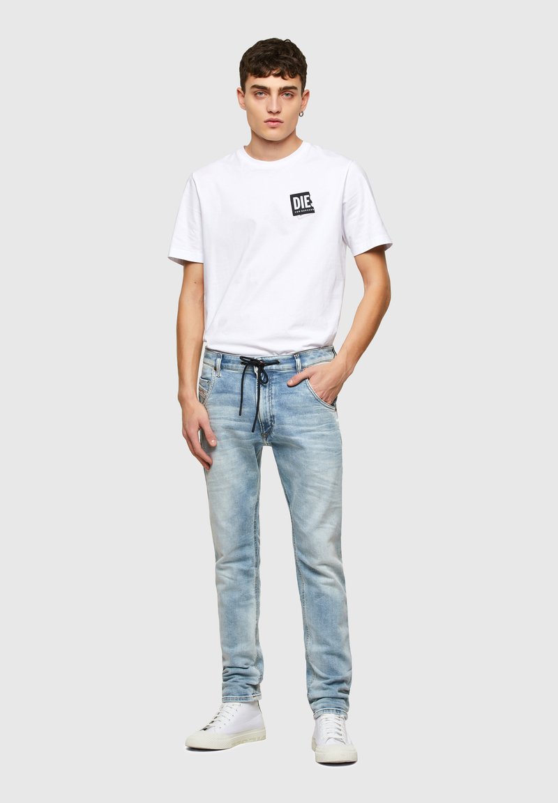 Weißes Baumwoll-T-Shirt mit einem schwarzen Grafikdesign in der oberen linken Ecke, kombiniert mit hellblauen Jeans mit Kordelzug und leichten Abnutzungen.