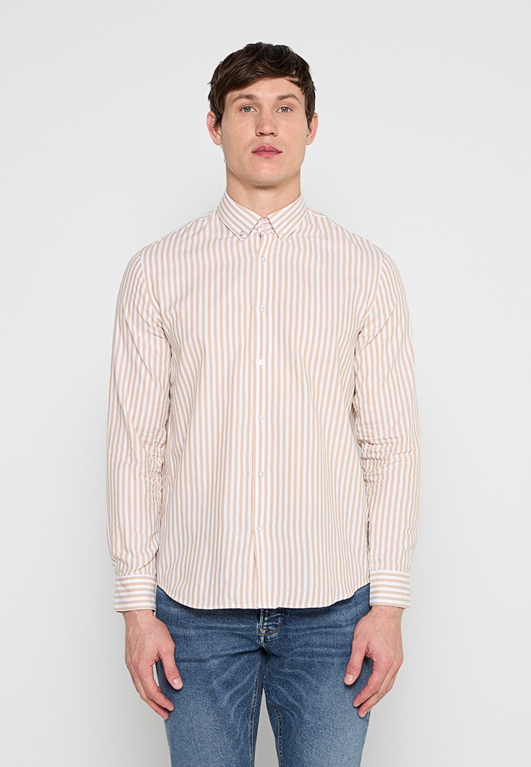 Mann trägt ein beige-weiß gestreiftes Hemd mit Knöpfen und Kragen sowie blaue Jeans, steht vor einem einfarbigen hellen Hintergrund.