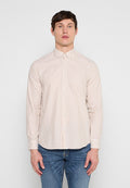 Mann trägt ein beige-weiß gestreiftes Hemd mit Knöpfen und Kragen sowie blaue Jeans, steht vor einem einfarbigen hellen Hintergrund.