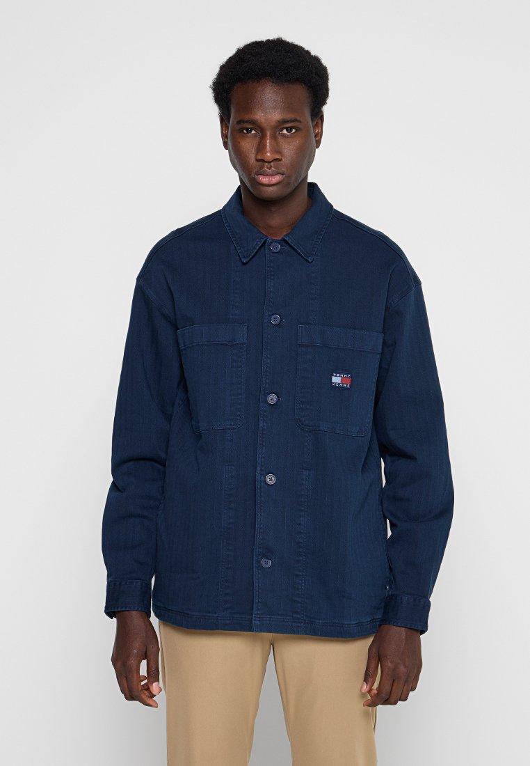 Junger Mann trägt eine dunkelblaue Jacke mit Knöpfen und Fronttaschen sowie beigen Hosen, steht vor einem schlichten hellen Hintergrund.