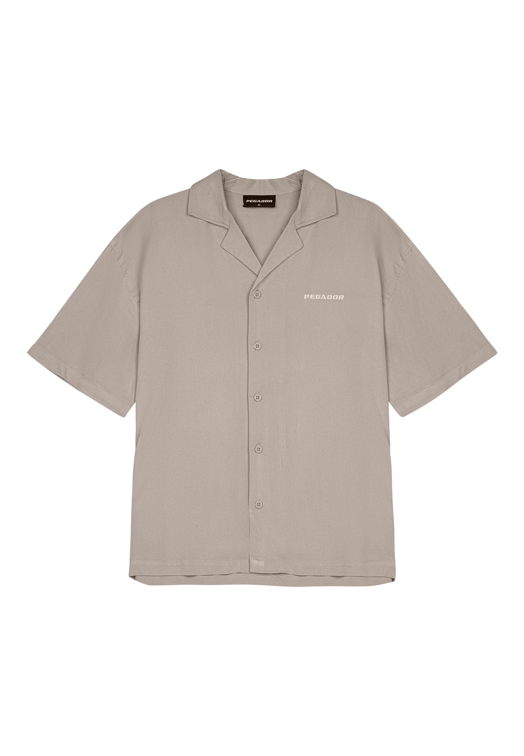Kurzärmliges, knöpfbares Hemd aus hellem Beige. Hat einen klassischen Kragen und einen dezenten "PEgador"-Druck auf der linken Brustseite.