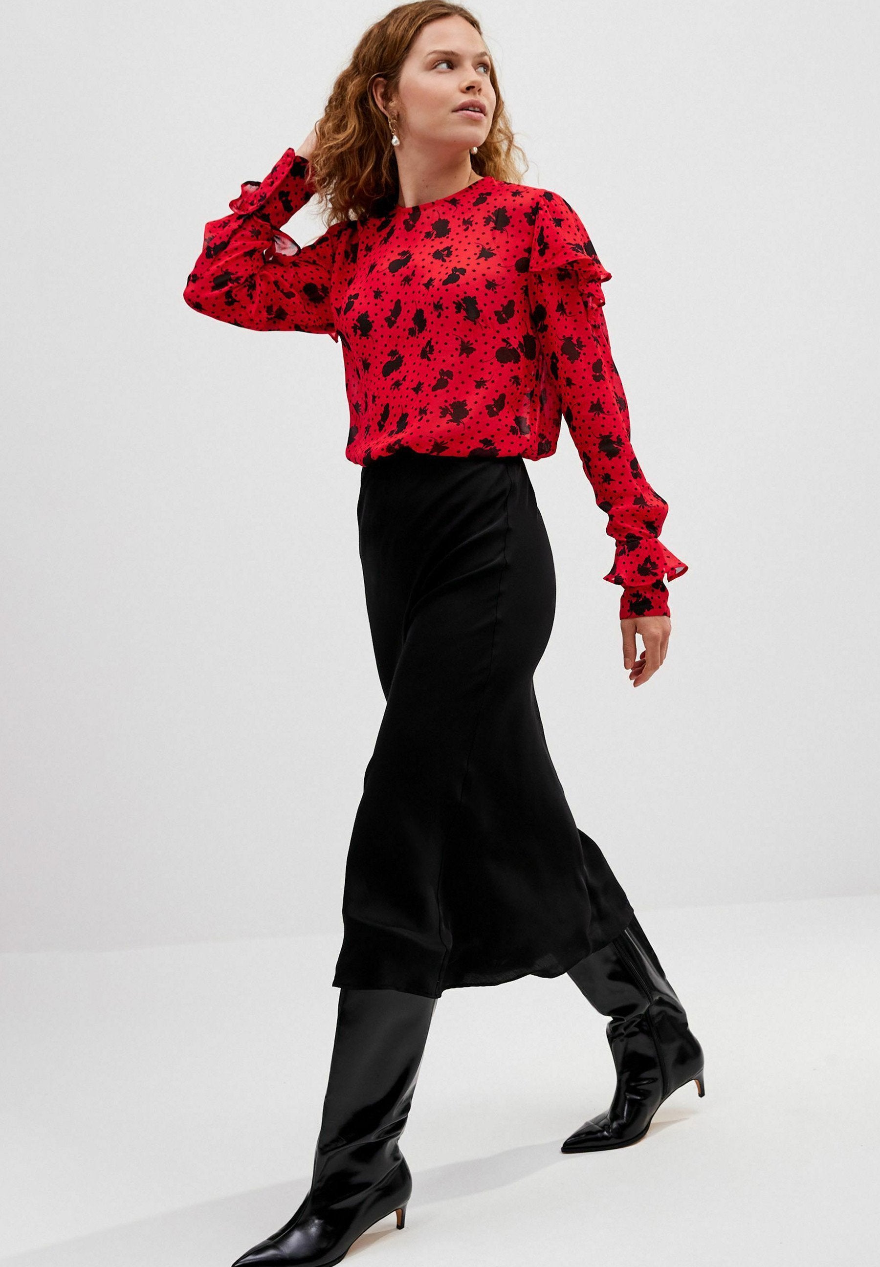 Rote Bluse mit schwarzem Spritzer-Muster und Rüschenschößen, kombiniert mit einem schwarzen Satin-Maxirock und knielangen schwarzen Stiefeln.