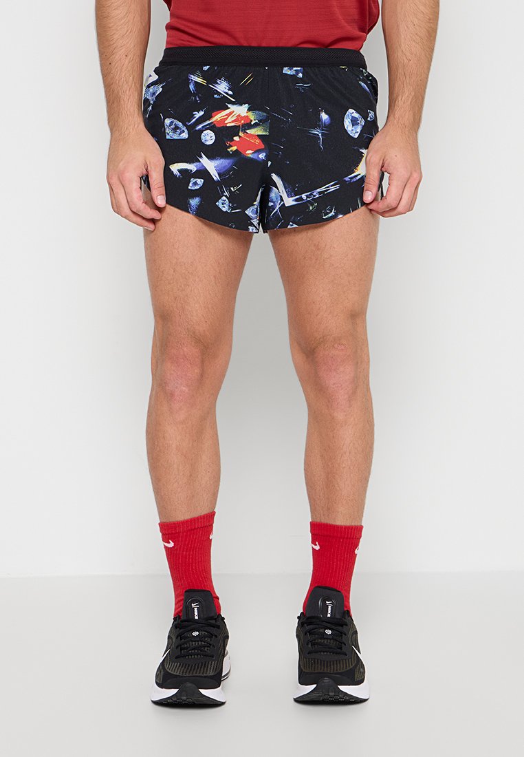 Mann trägt schwarze Sportshorts mit bunten abstrakten Mustern, rote Sportsocken, schwarze Laufschuhe und ein rotes Shirt, steht vor hellem Hintergrund.