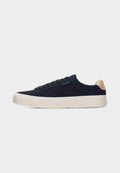 Niedrig geschnittener Sneaker in Marineblau mit beigem Fersenlasche, weißer dicker Sohle und Schnürverschluss, von der linken Seite auf hellem grauem Hintergrund dargestellt.
