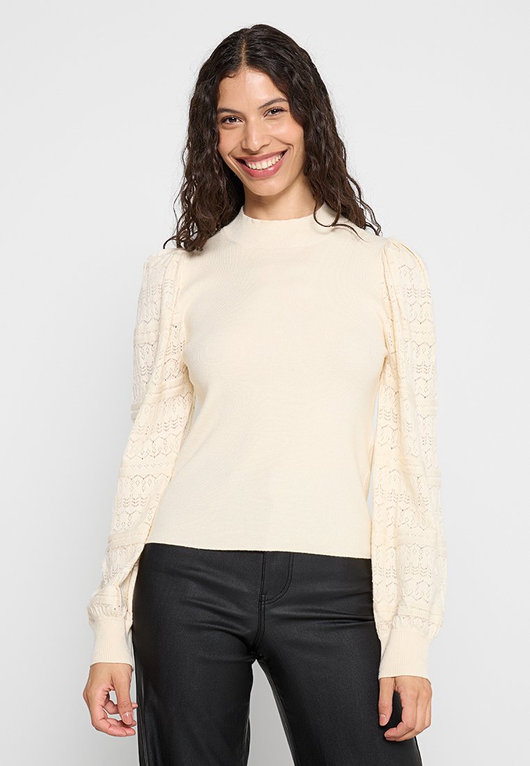 Cremesweater mit geripptem Körper und Puffärmeln im Spitzenmuster. Verfügt über einen hohen Kragen und enganliegende Bündchen, kombiniert mit schwarzen Hosen.