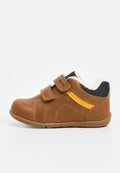 Braune Kindersneaker mit zwei Klettverschlüssen, gelbem seitlichem Akzent, schwarzem gepolstertem Kragen und weißem Fleece-Futter, flache Sohle auf weißem Hintergrund.
