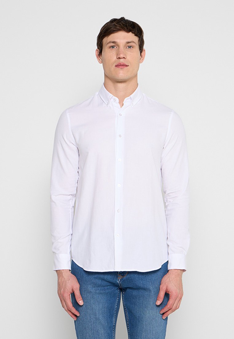 Ein Mann steht vor einem einfachen hellen Hintergrund, trägt ein weißes, geknöpftes Langarmhemd und blaue Jeans.