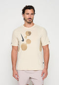 Mann mit dunklem Haar und Bart trägt beige Nike-T-Shirt mit abstrakten braunen Formen und passende helle Shorts, stehend vor einfarbigem Hintergrund.