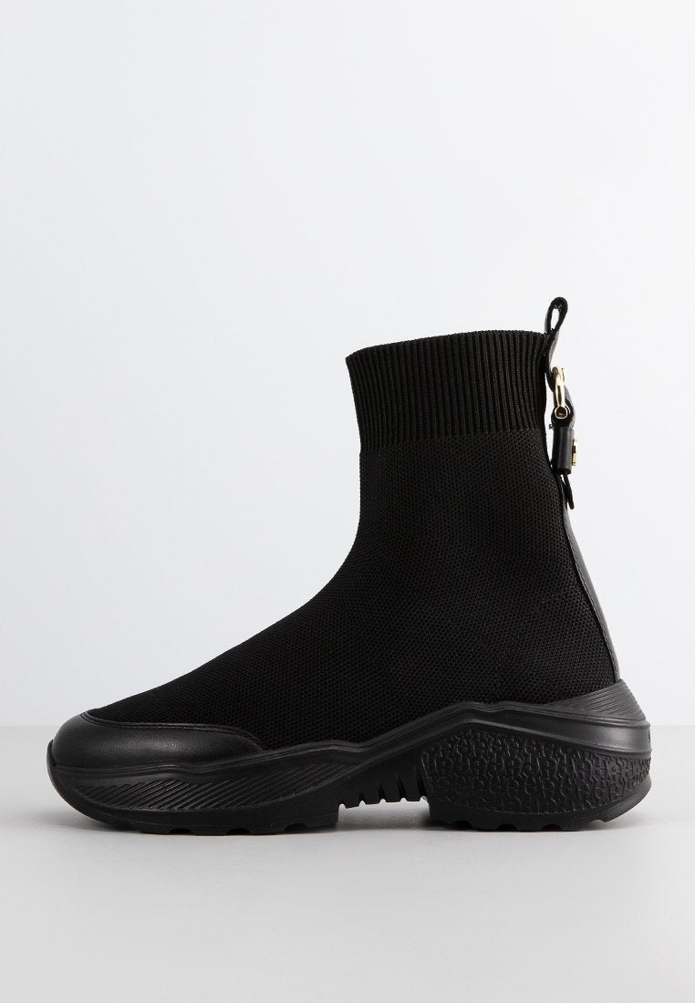 Schwarze Knöchelstiefel aus dehnbarem Strickmaterial mit strukturiertem Sohlen, glattem Zehenbereich und einer Schlaufe mit goldfarbener Hardware.