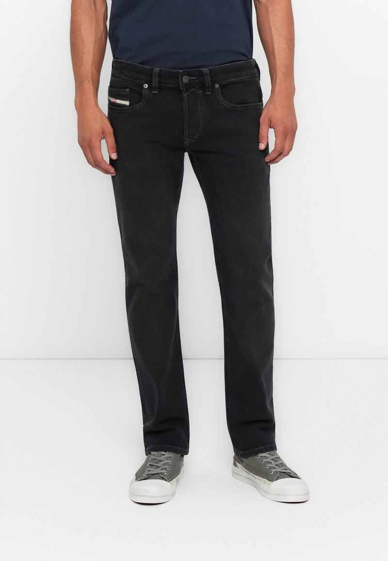 Schwarze Denim-Jeans im Slim-Fit, mit Fünf-Taschen-Design und sichtbaren Nähten. Kombiniert mit grauen Sneakers, vor einem weißen Hintergrund.