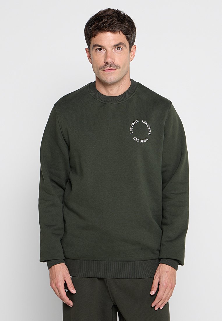 Dunkelgrüner Pullover mit einem runden weißen Logo auf der Brust, Rundhalsausschnitt, langen Ärmeln und gerippten Bündchen und Saum. Weiche Textur.