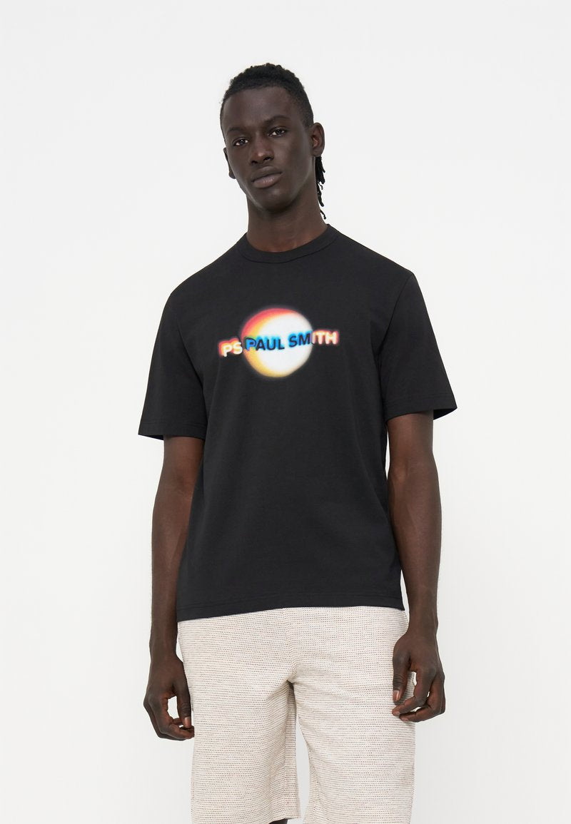 Mann trägt schwarzes T-Shirt mit buntem, unscharfem "PS Paul Smith"-Logo und hell strukturierten Shorts, steht vor einfarbig weißem Hintergrund.