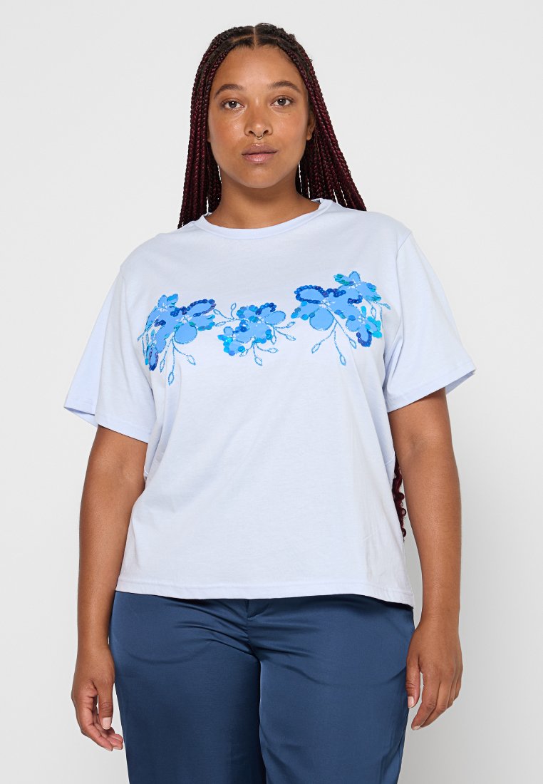 Eine Frau mit langen geflochtenen Haaren trägt ein weißes T-Shirt mit blauem, floralen Paillettenmuster und marineblauen Hosen und steht vor einem einfarbigen Hintergrund.