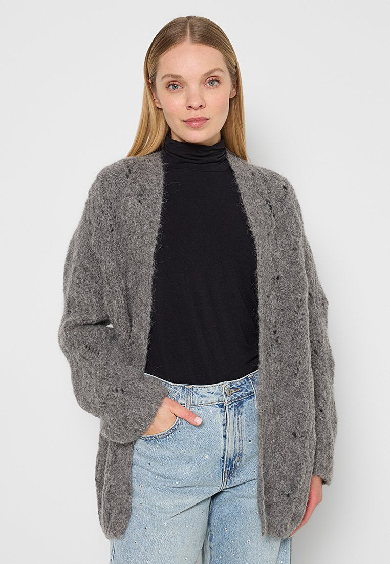 Frau trägt eine flauschige graue Strickjacke, einen schwarzen Rollkragenpullover und hellblaue Jeans mit kleinen Strasssteindetails, steht vor einem schlichten Hintergrund.