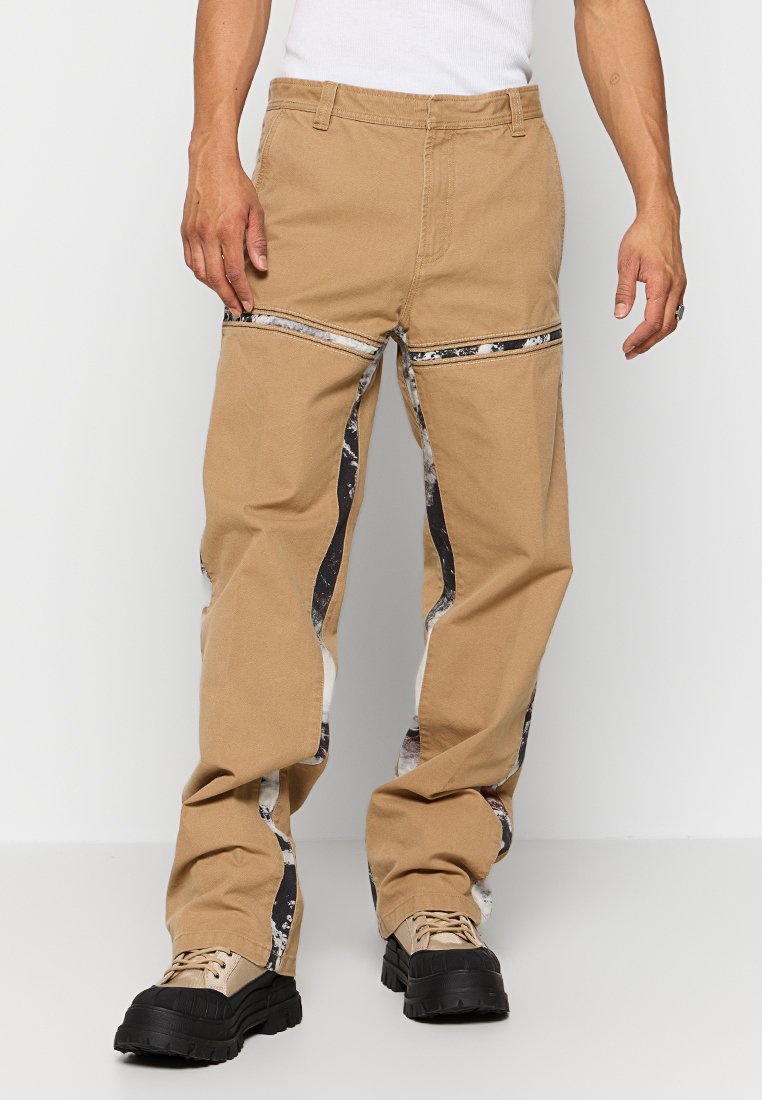 Beige weit geschnittene Hose mit abstrakten schwarzen und weißen Seitenpaneelen, ausgestattet mit einem Reißverschluss und einem robusten Stoff. Texturiert mit einer glatten Oberfläche.