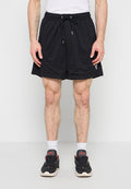 Mann trägt schwarze Sportshorts mit Kordelzug, weiße Socken und schwarze Turnschuhe, steht vor einem schlichten Hintergrund.